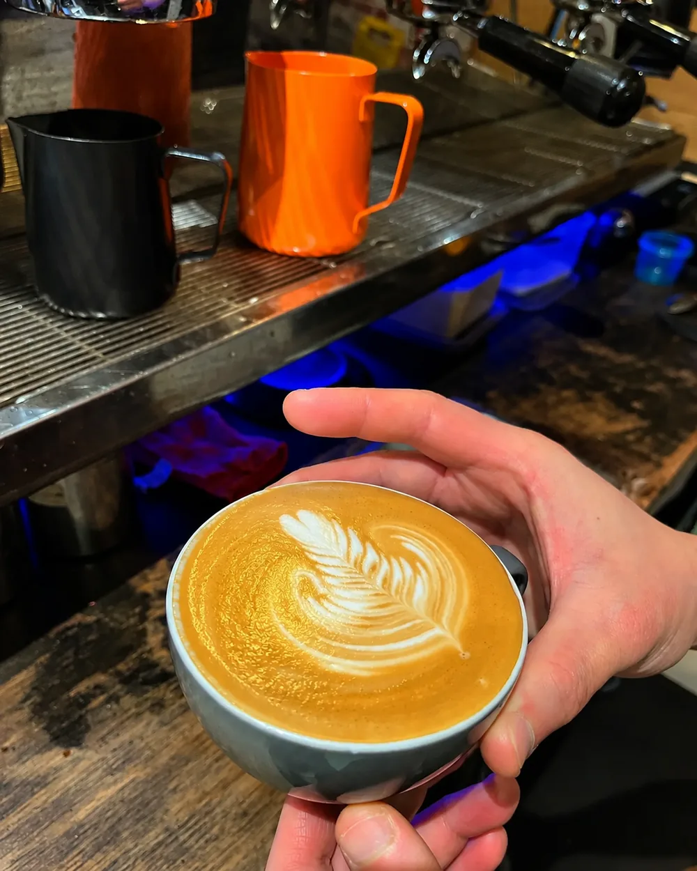 A cup of coffee with latte art held beside N’Joy’s espresso machine